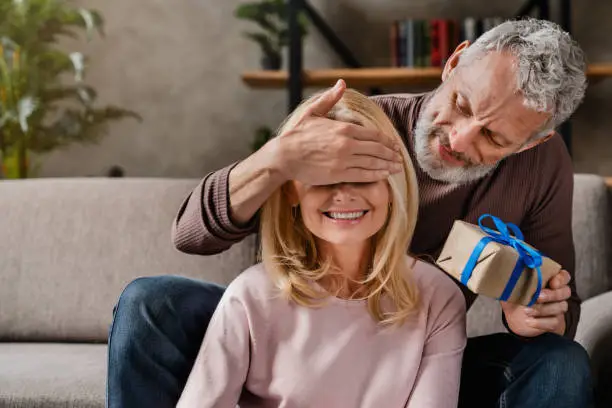 Photo of Middle aged man making surprise with gift box to his wife closing her eyes while celebrating anniversary or birthday at home