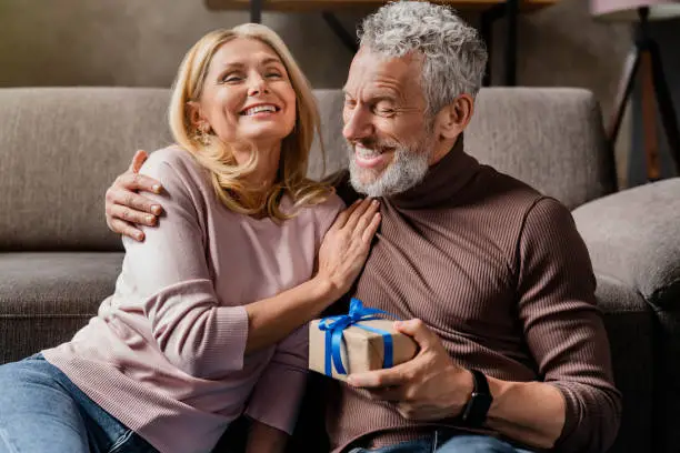 Photo of Loving middle aged man making surprise by giving gift in box to his wife celebrating anniversary or birthday at home