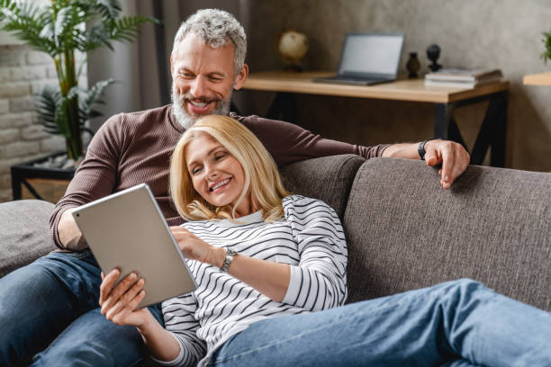 glückliches senior-paar mit digitalen tablet auf sofa in innenräumen - senior adult domestic life cheerful female stock-fotos und bilder