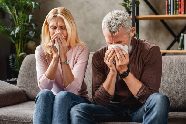 coppia di mezza età seduta sul divano che soffia il naso, concetto influenzale con tessuti e coperta - cold and flu flu virus bed couple foto e immagini stock