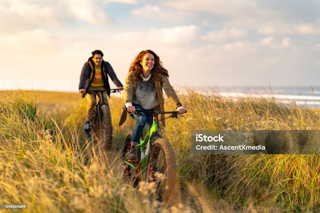 Young couple ride fat bikes on coastal trail Sun lights up surrounding grassy meadow, Pacific Ocean in distance Cycling Stock Photo