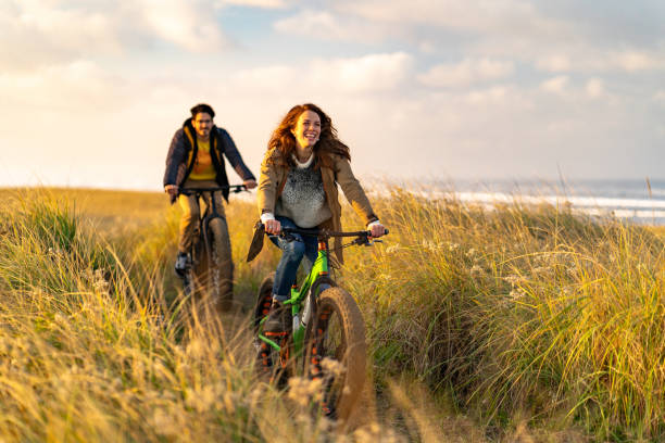 Young couple ride fat bikes on coastal trail Sun lights up surrounding grassy meadow, Pacific Ocean in distance travel lifestyle stock pictures, royalty-free photos & images