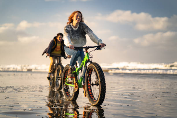 Young couple ride fat bikes on the beach, tidal flat Waves roll in as they ride through the shallows pacific northwest photos stock pictures, royalty-free photos & images