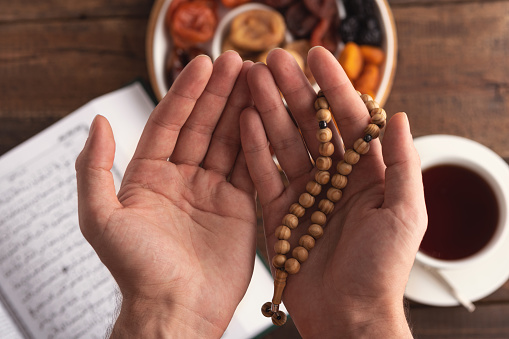 top view men hand of prayer with wooden beads in sunlight, iftar concept, Ramadan month, Koran, plate of dried fruit, Cup of tea on wooden table