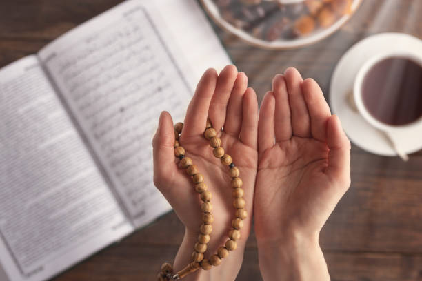 top view female hand of prayer with wooden beads in sunlight, iftar concept, Ramadan month, Koran, plate of dried fruit, Cup of tea on wooden table female hand of prayer with wooden beads in sunlight, iftar concept, Ramadan month, Koran, plate of dried fruit, Cup of tea on wooden table allah photos stock pictures, royalty-free photos & images