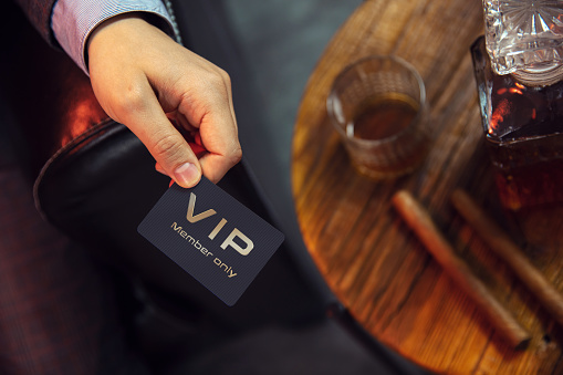 El hombre tiene la tarjeta de miembro VIP. Vista desde la parte superior de la mano del caballero que sostiene una tarjeta de membresía VIP exclusiva junto a la mesa de madera con whisky en jarra y vaso con puros. photo