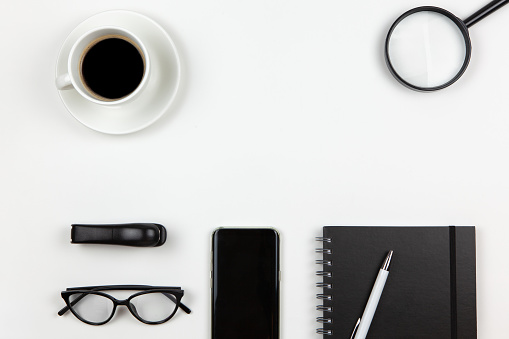 Modern unisex working space, top view. Notepad, pen, coffee, smart phone, magnifier, glasses on white background, copy space, flat lay. Desktop of freelancer. Work from home concept. Monochrome image.