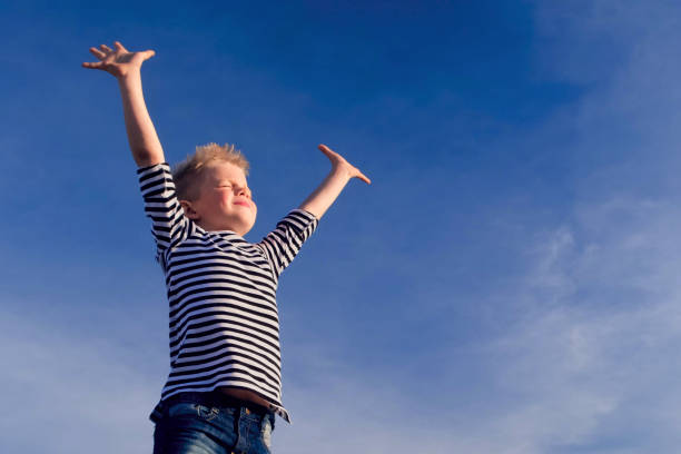 Relaxed boy breathing fresh air raising arms over blue sky at summer. Dreaming, freedom and traveling concept stock photo