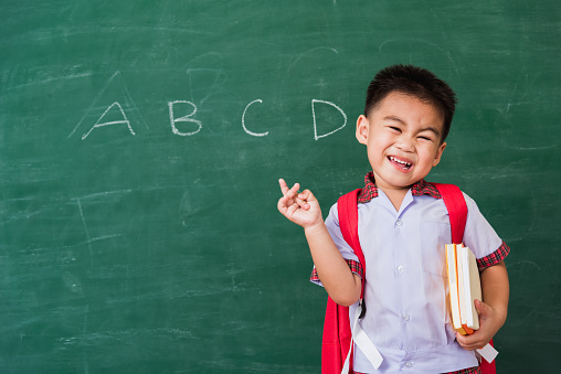 Back to School. Happy Asian funny cute little child boy kindergarten in student uniform and school bag and books smile point finger space side away on school blackboard, First time to school education