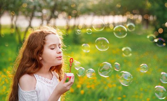Beautiful girl blowing soap bubbles in summer park. Happy childhood concept.