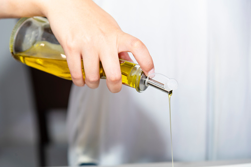 Close up of a female hand holding an olive oil dispenser with oil pouring out of it on an out of focus background. Healthy food concept.