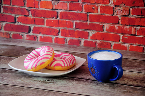 A plate with two donuts in pink strawberry glaze and a blue ceramic cup with cappuccino is on a wooden table against a red brick wall. Close up.