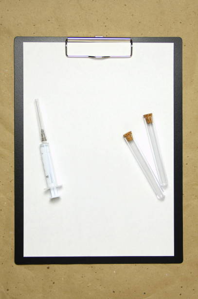 The tablet with a clip for the clip of paper with a white sheet a4 lies against the background of craft brown crumpled paper. On top is a medical syringe and two transparent empty tubes with stoppers. Place for text and layout for design. la tableta con un clip para el clip de papel con una hoja blanca a4 se encuentra sobre el fondo del papel crujiente marrón artesanal. en la parte superior hay una jeringa médica y dos tubos vacíos transparentes con tapones. lugar para texto, espacio de
