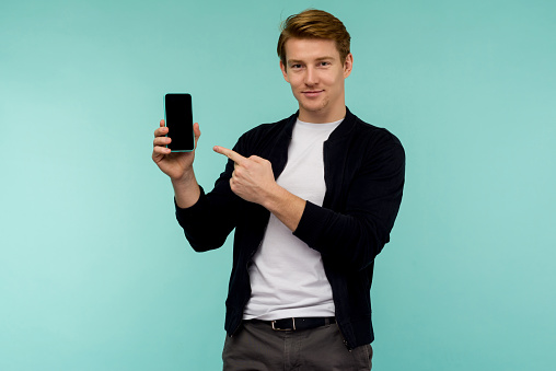 Cheerful sporty red-haired guy shows a finger on the smartphone screen on a blue background. - image