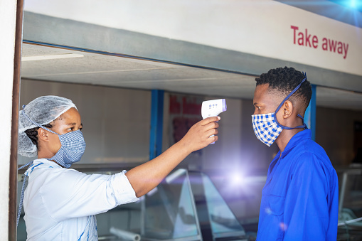 African woman working at a shop, using a mask for protection, registering and checking temperature of the customers with an infrared thermometer