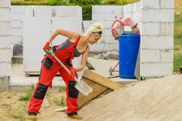 Woman working hard on construction site, using shovel digging sand soil. Partially built new home early stage. Industry.