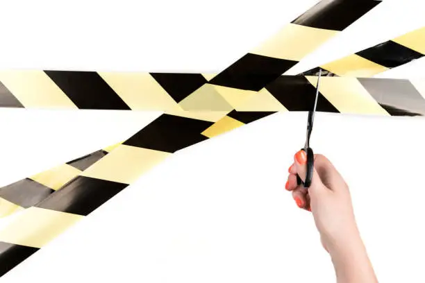 Yellow-black restrictive tape on a light background. Female hand holds scissors to cut the tape. End quarantine and remove restrictions