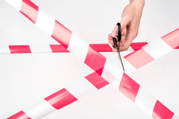 Red-white restrictive tape on a light background. Male hand holds scissors to cut the tape.