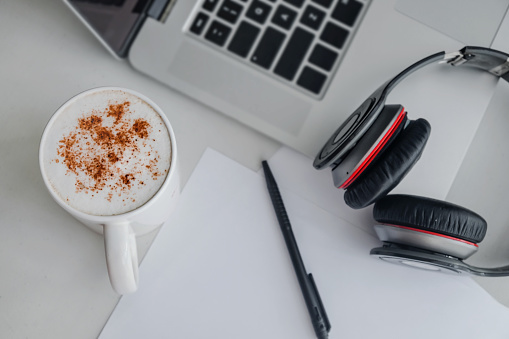Close up shot of business objects for morning conference call above desk table which consists of  headphones , blank paper ballpoint pen and hot mochaccino, next to laptop in home office