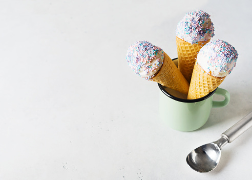 White vanilla ice cream scoop in waffle cone with sprinkles in cup on light grey background, top view, copy space