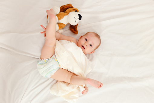 Baby less than a year lies on the bed with a soft toy. Baby in diaper lifts legs up