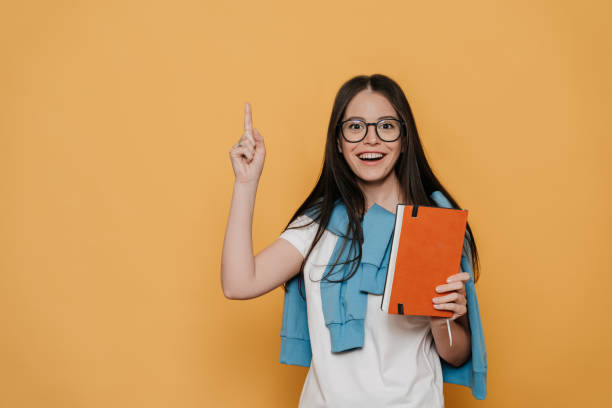 istock Great idea. Young cheerful brunette in glasses dressed in casual, holds her diary, pointing upward in good  idea inspiration, has amazed face, over yellow backdrop. Education people concept. 1248459781