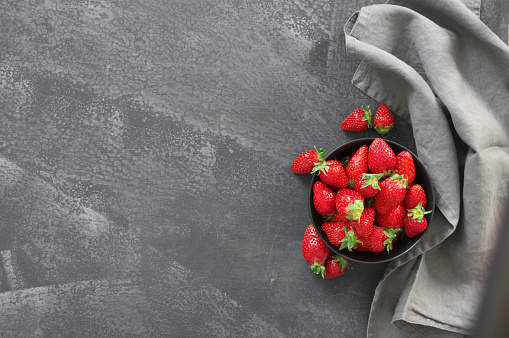 Pile of fresh strawberries in black ceramic bowl with linen napkin on grey rough background. Top view.