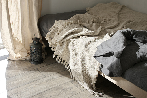 Unmade bed with cotton boho blanket in cozy simple bedroom at sunlight. Good morning concept.