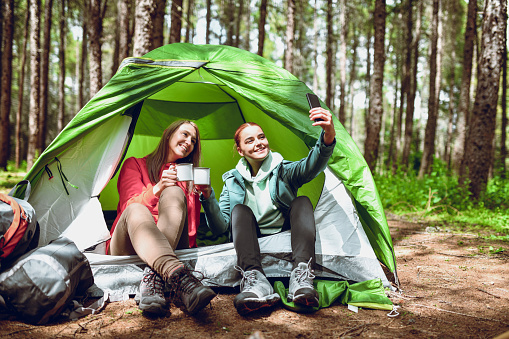 Cute Female Campers Taking Selfie While Drinking Tea And Sitting In Front Of Tent