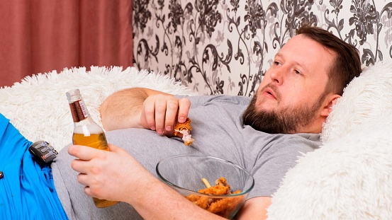 fat man drinks beer and eats unhealthy food chicken wings, bored in front of the TV outlook on the couch. The concept of malnutrition, quarantine at home, alcoholism