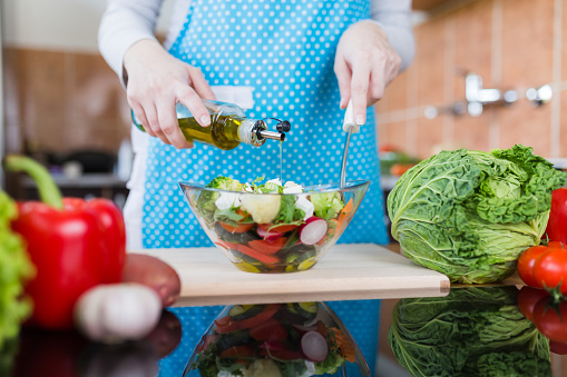 Close up of woman in apron preparing vegetable salad and adding olive oil at home kitchen desk.