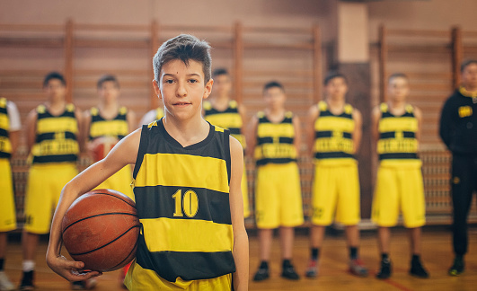 Group of young teenage boys, basketball players, standing in sports hall with their coach, one teenage boy standing in front and looking at camera.