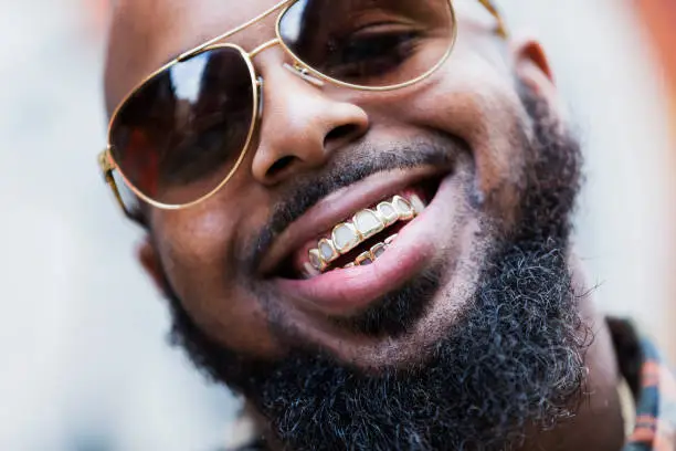 An African-American man with a gold grill, thick beard and sunglasses, smiling.