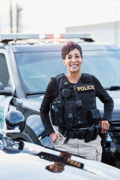 African-American policewoman standing by patrol car An African-American policewoman standing next to her patrol car, smiling confidently at the camera. She is a mature woman in her 40s, wearing a bulletproof vest and duty belt. female police officer stock pictures, royalty-free photos & images