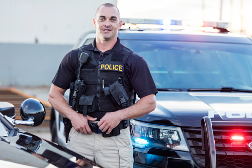 A policeman standing next to his patrol car, looking at the camera, smiling. He is a mid adult m an in his 30s, wearing a bulletproof vest and duty belt.