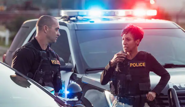 Two multi-ethnic police officers standing by the patrol cars, wearing bulletproof vests and duty belts. The policewoman is a mature African-American woman in her 40s. Her partner is a mid adult man in his 30s. She is talking into her radio.