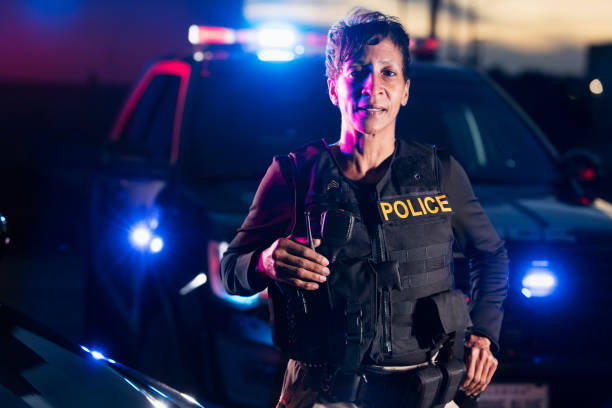 African-American policewoman standing by patrol car An African-American policewoman standing next to her patrol car, looking at the camera with a serious but confident expression on her face. She is a mature woman in her 40s, wearing a bulletproof vest and duty belt. It is nighttime and the emergency lights on her patrol car are on. female police officer stock pictures, royalty-free photos & images