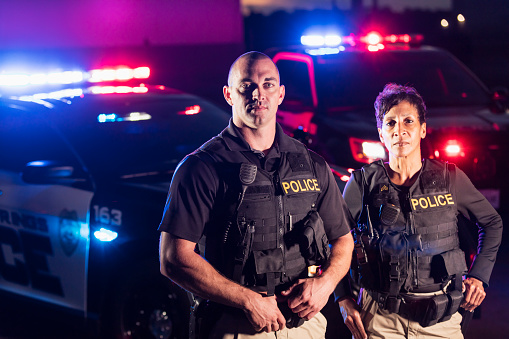 Two multi-ethnic police officers standing in front of patrol cars, wearing bulletproof vests and duty belts. The policewoman is a mature African-American woman in her 40s. Her partner is a mid adult man in his 30s. It is nighttime and the emergency lights on top of the vehicles are flashing.