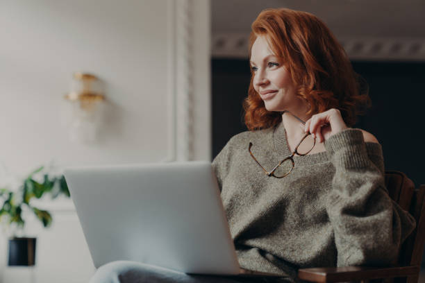 istock Experienced redhead female entrepreneur makes plans, looks aside with dreamy expression, uses modern technology, holds spectacles, wears casual jumper, enjoys distance education and e learning 1248519463