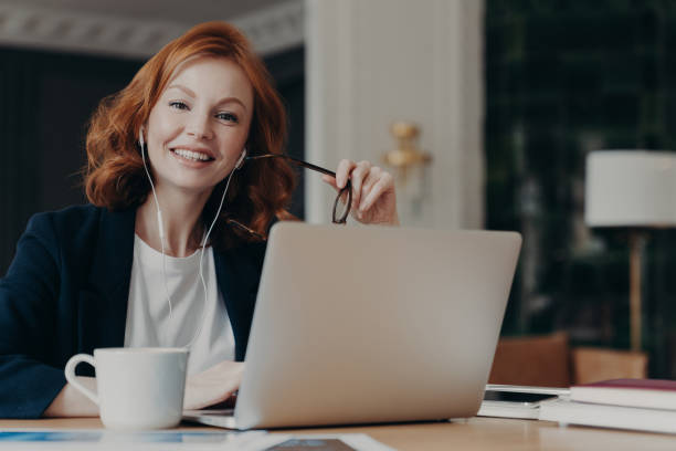 istock Young happy female freelancer with ginger hair thinks about new ideas for work, works distantly on laptop computer, prepares for video conference, uses earphones, poses at workplace with hot drink 1248519504