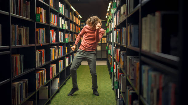 University Library: Handsome Caucasian Student Celebrates Successful Pass of Exams, Dances Between Rows of Bookshelves. Success in College: Admission, Graduations, Finishing Master Thesis University Library: Handsome Caucasian Student Celebrates Successful Pass of Exams, Dances Between Rows of Bookshelves. Success in College: Admission, Graduations, Finishing Master Thesis dissertation photos stock pictures, royalty-free photos & images