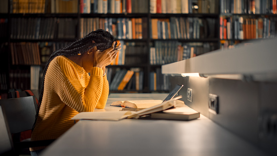 University Library: Exhausted and Tired Black Girl uses Laptop, Writes Essay, Study for Class Assignment. Students Learning, Studying for Exams College. Side View Portrait with Bookshelves