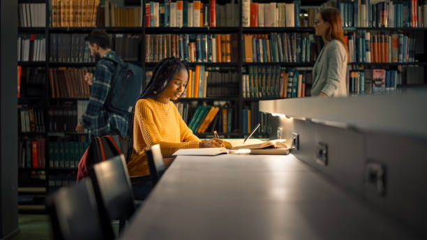 universiteitsbibliotheek: begaafd zwart meisje maakt gebruik van laptop, schrijft notities voor het papier, essay, studie voor klasse opdracht. studenten leren, studeren voor examens college. zijaanzichtportret met boekenplanken - library stockfoto's en -beelden
