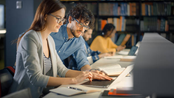 istock University Library: Talented Caucasian Girl uses Laptop, Smart Helpful Classmate Explains and Advices Her with Class Assignment. Happy Diverse Students Talking, Learning, Studying Together for Exams 1248521817