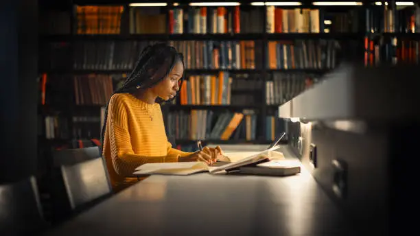 University Library: Gifted Black Girl uses Laptop, Writes Notes for the Paper, Essay, Study for Class Assignment. Students Learning, Studying for Exams College. Side View Portrait with Bookshelves