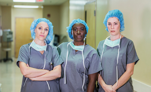 A multi-ethnic group of three female doctors or nurses standing in a hospital corridor, wearing scrubs and surgical caps. The team of healthcare works are staring at the camera with serious expressions.