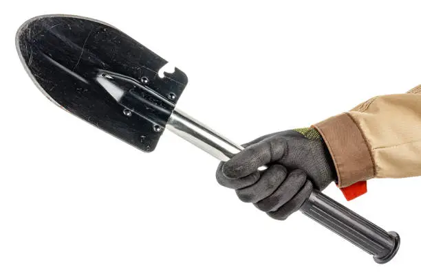 Black folding shovel with with scratches and scuffs in male hand in black protective glove and brown uniform isolated on white background