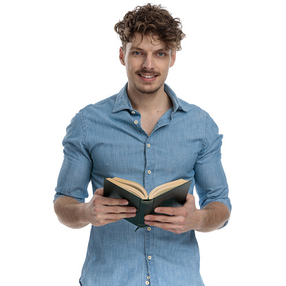 smiling young casual guy in denim shirt reading book and standing isolated on white background, portrait
