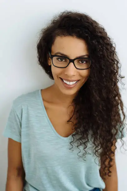 Young woman wearing glasses with a friendly smile and long frizzy black hair looking at the camera with a happy expression against a white studio background