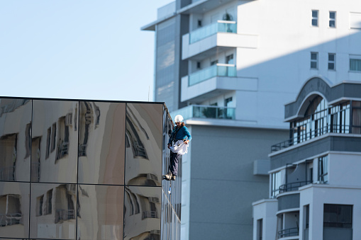 Image of a young skyscraper window washer wearing safety equipments and cleaning a glass curtain wall building facade